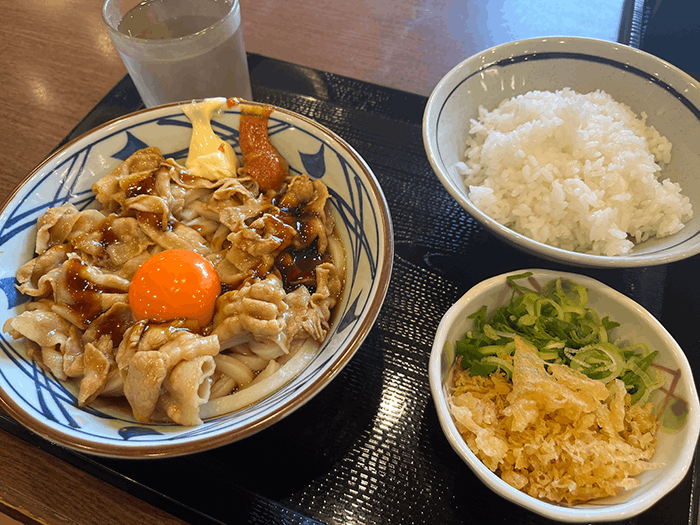 丸亀製麺こく旨豚玉ぶっかけと白ご飯、天かすなどのトッピングが写った画像